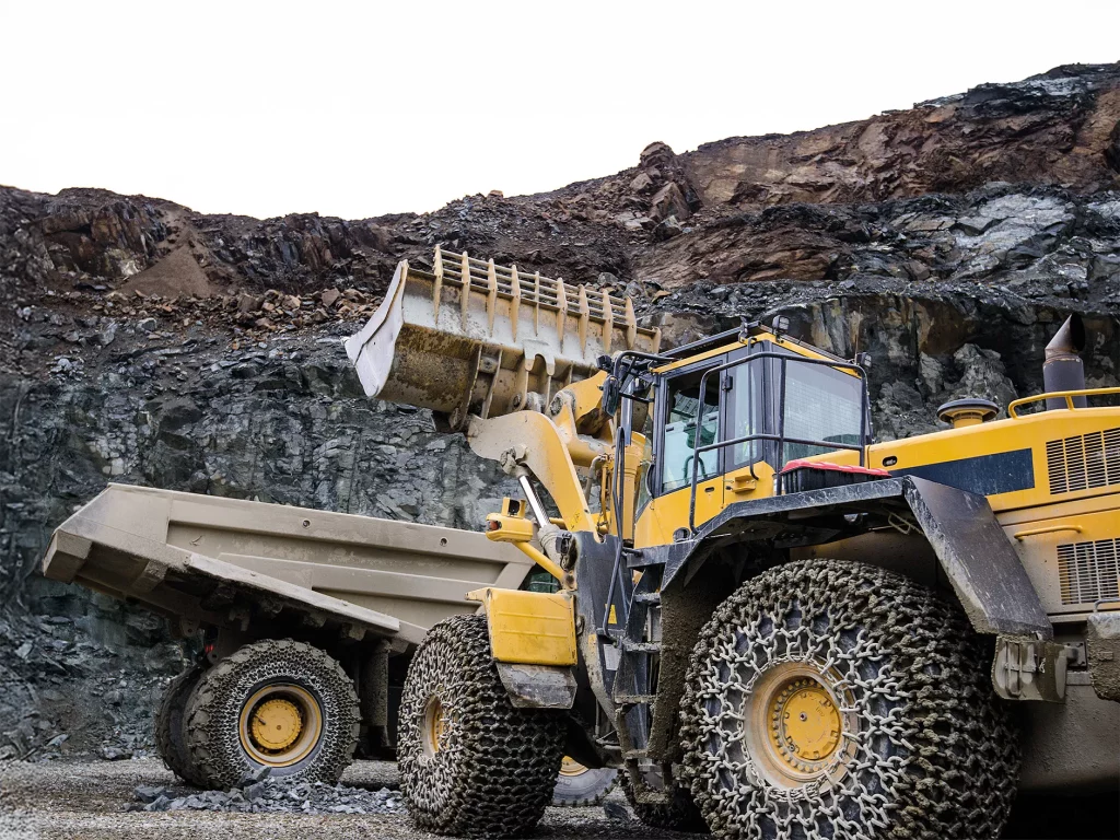 Two large scale earth moving machines working in a stone quarry setting. Both vehicles are fitted with pewag tire protection chains or TPC