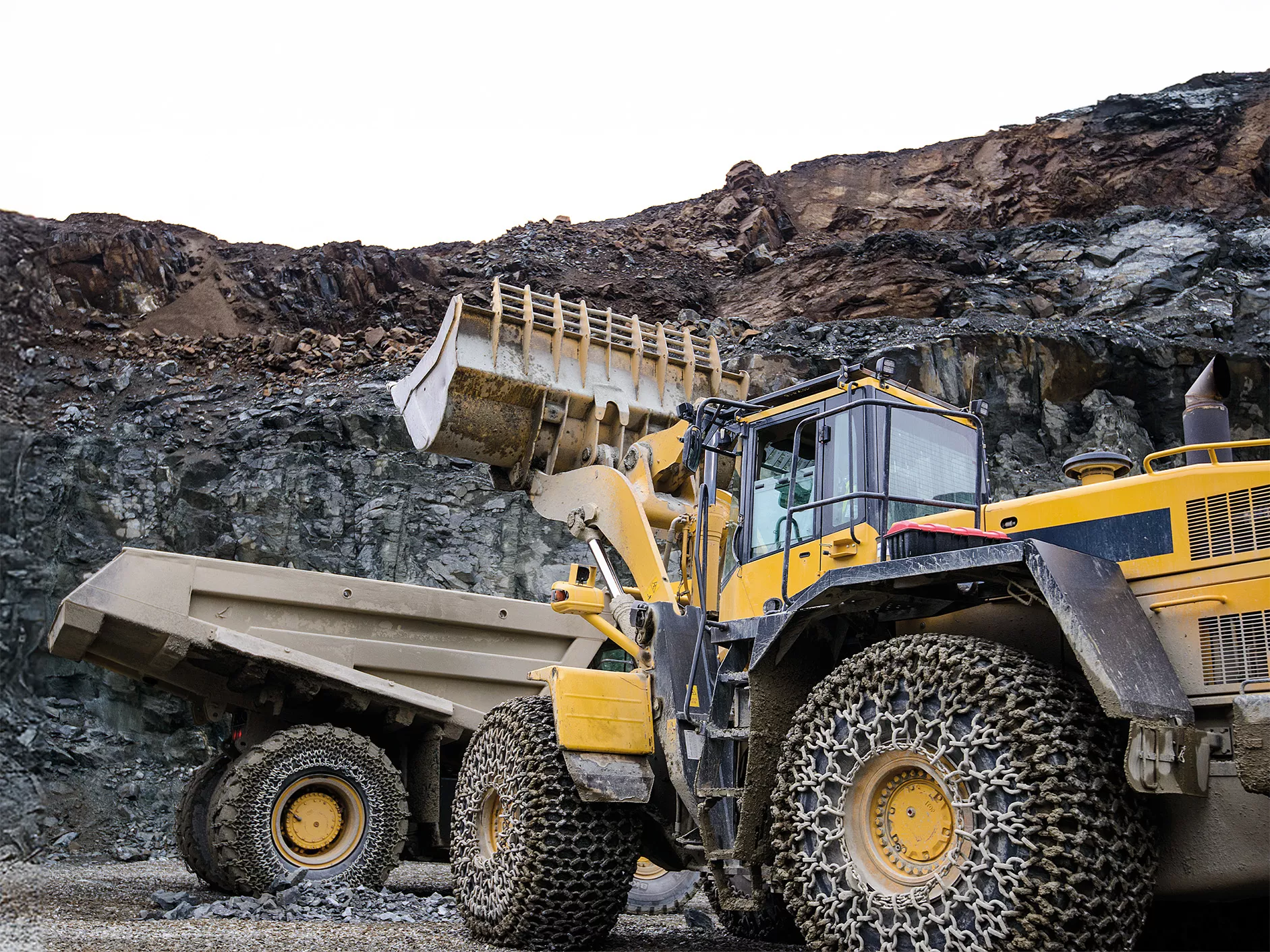 Two large scale earth moving machines working in a stone quarry setting. Both vehicles are fitted with pewag tire protection chains or TPC